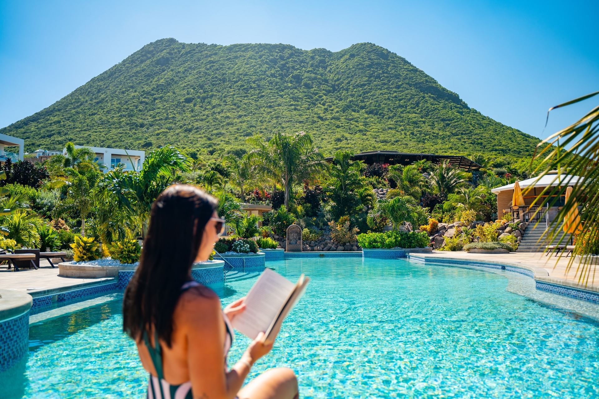 Dame ontspant bij Hummingbird Pool en leest een boek, omringd door groen en een schilderachtige berg bij Golden Rock Resort