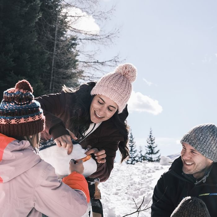Familie baut Schneemann, Mutter und Kind formen Kugel, Vater beobachtet im Schnee mit Bäumen im Hintergrund.