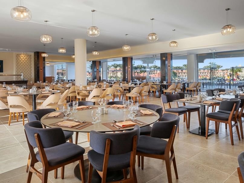 An elegant restaurant with round tables, chairs, and large windows offering a view of the outdoors.