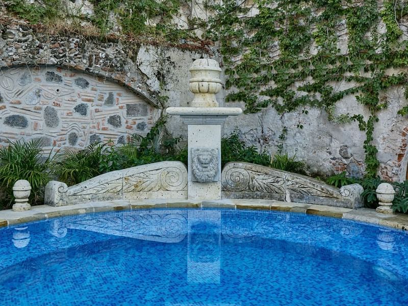 Stone fountain at Fiesta Americana, surrounded by lush greenery and an ivy-covered wall, complementing a blue tiled pool