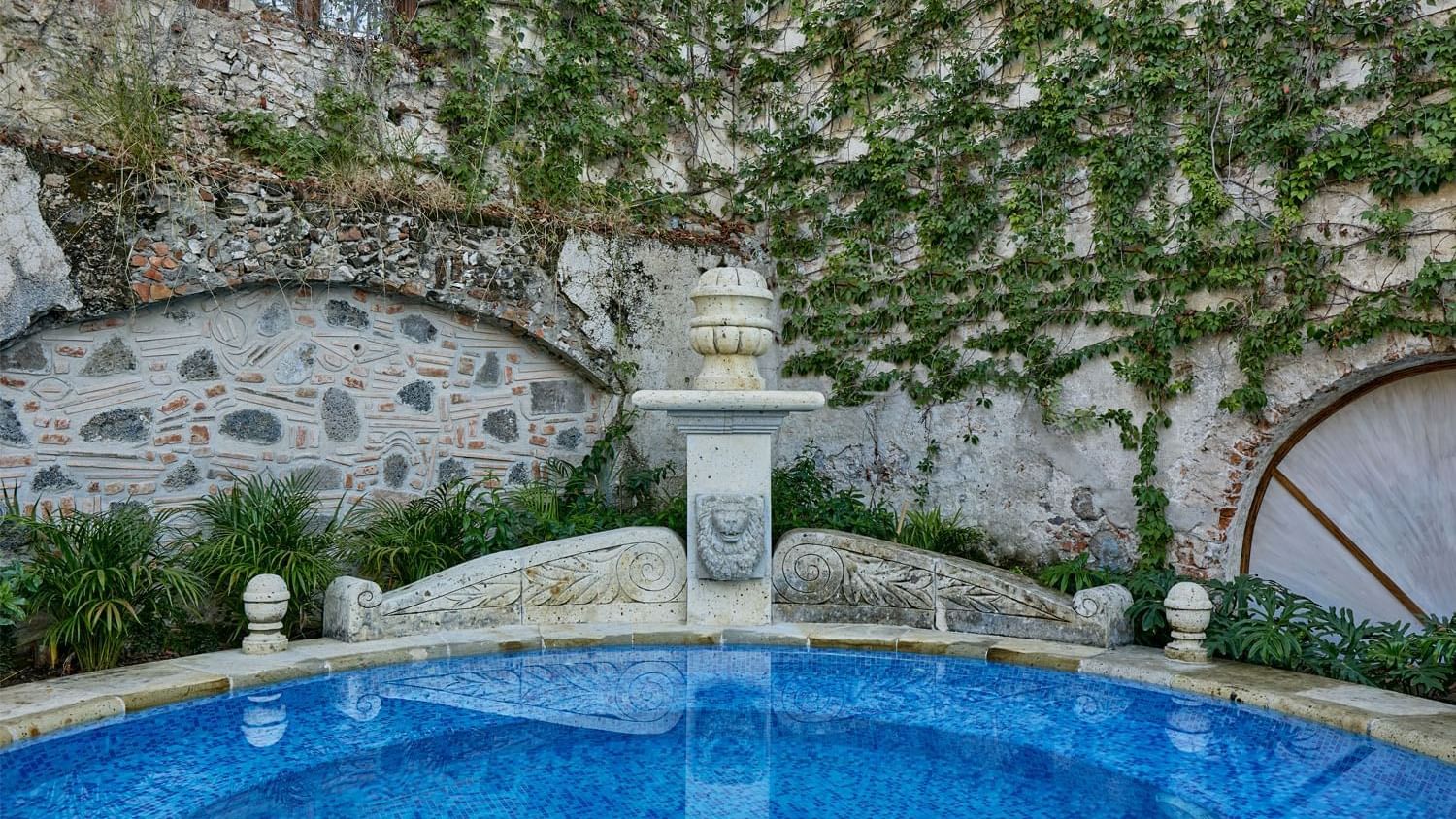 Stone fountain at Fiesta Americana, surrounded by lush greenery and an ivy-covered wall, complementing a blue tiled pool