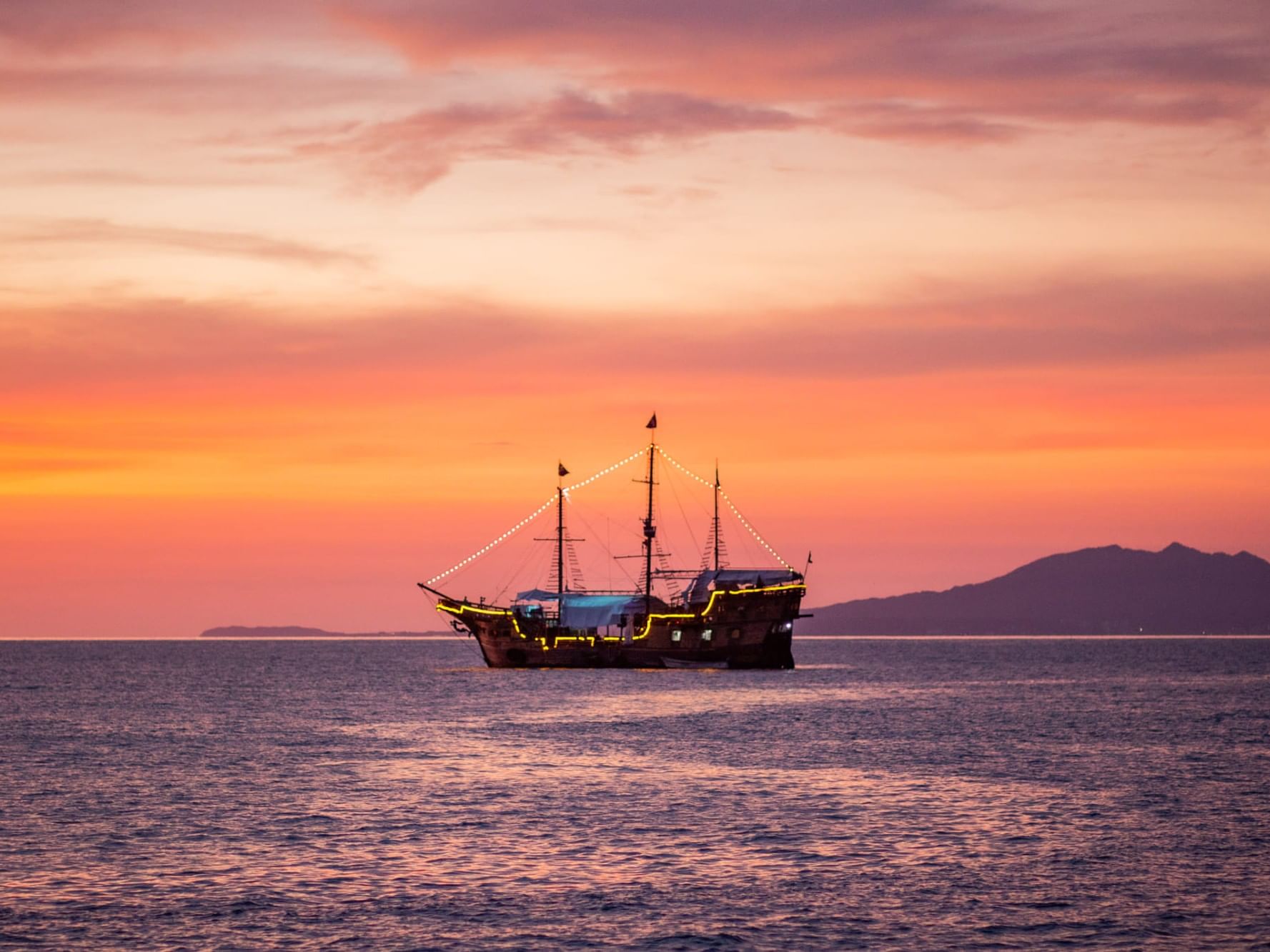 Barco pirata navegando en el mar al atardecer con cielo rosado y montañas al fondo, Marigalante II en Vallarta