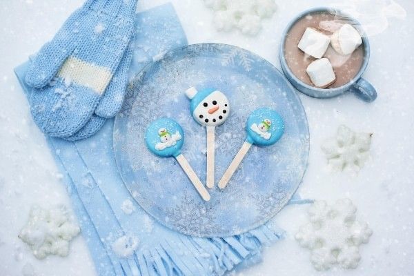 A plate of snowman-themed desserts with hot chocolate, blue mittens, and a blue scarf resting on a bed of snow.