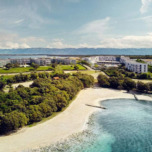 Luftaufnahme eines Strandes mit weißen Gebäuden und Bergen im Hintergrund.