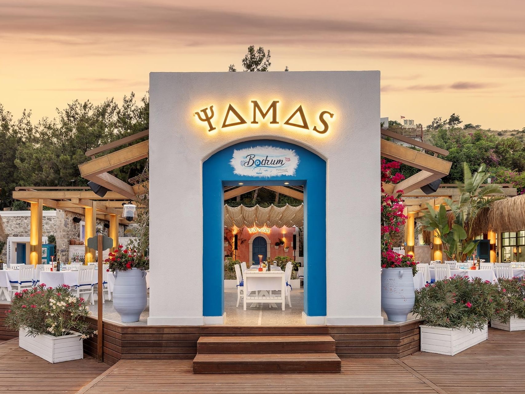 Chic restaurant entrance at Titanic Luxury Collection Bodrum, with a blue door, floral décor, and illuminated signage.