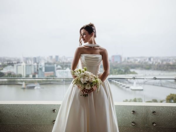 Bride in white wedding dress holding bouquet of flowers, on balcony overlooking Brisbane River at Emporium Hotel