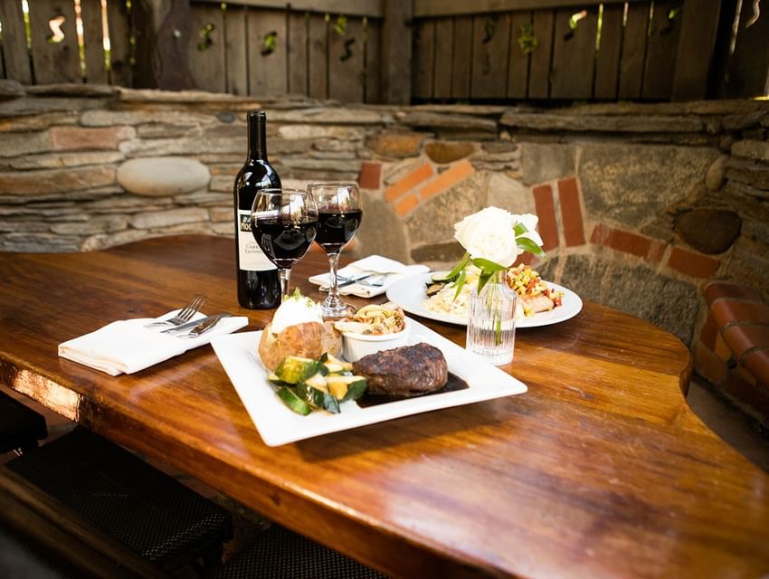Two plates of food, wine, and utensils on a wooden table against a stone wall.