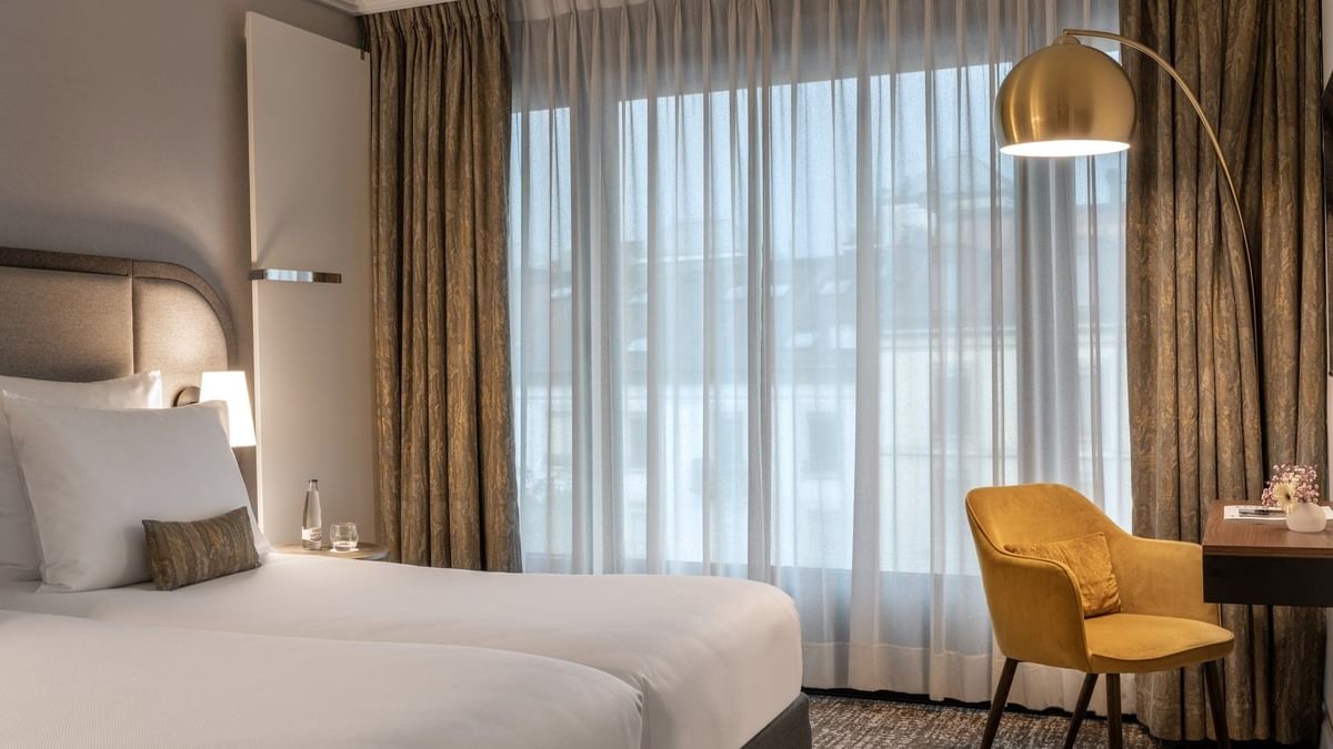 Twin beds by a yellow velvet armchair under a gold arched floor lamp near drapes in Classic Room at Warwick Geneva