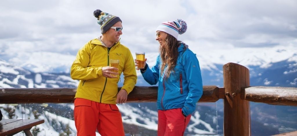 Couple enjoying drinks on a mountain patio overlooking snowy Whistler peaks.