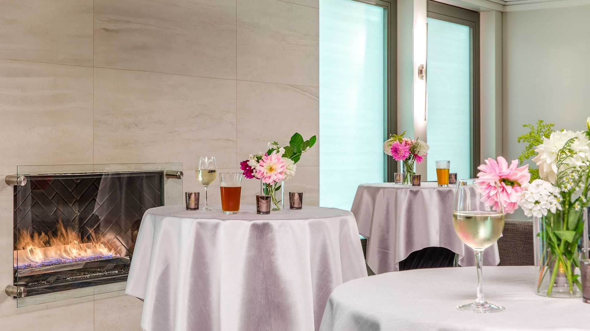 A cozy lounge area with a fireplace, round tables with white tablecloths, flowers, and glasses.