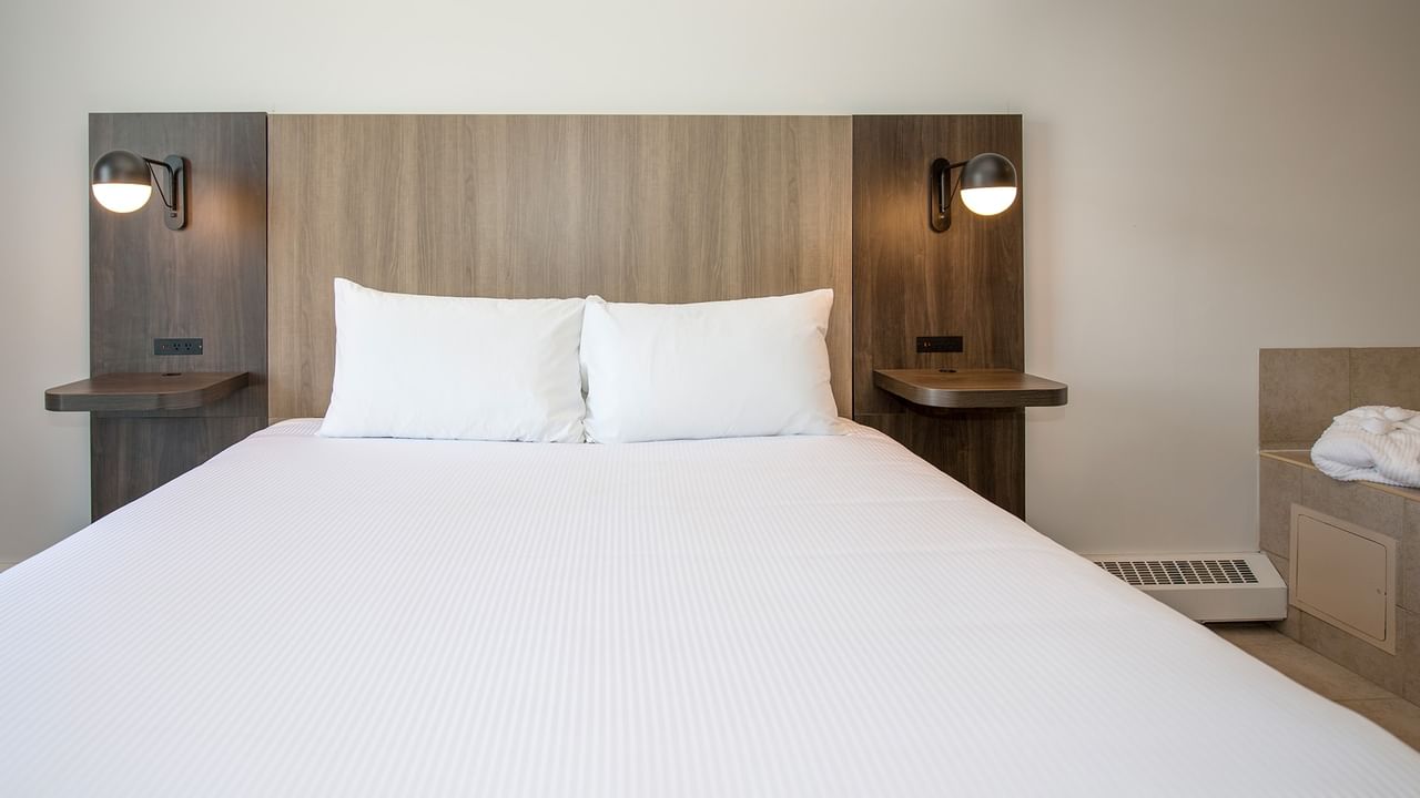 A white bed with two pillows, wooden headboard, and wall-mounted lights on either side.