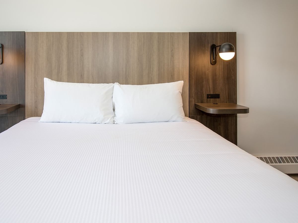 A neatly made bed with two white pillows, wooden headboard, and wall-mounted lamps on either side.