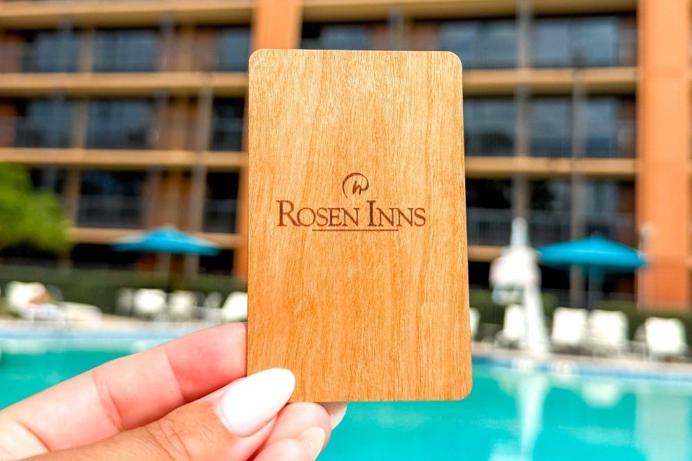 A close-up of a hand holding a wooden Rosen Inns key card against a backdrop of the hotel and pool in Orlando.