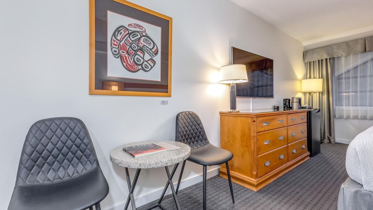 Hotel room with two chairs, a small table, a dresser with a TV, and a framed artwork.