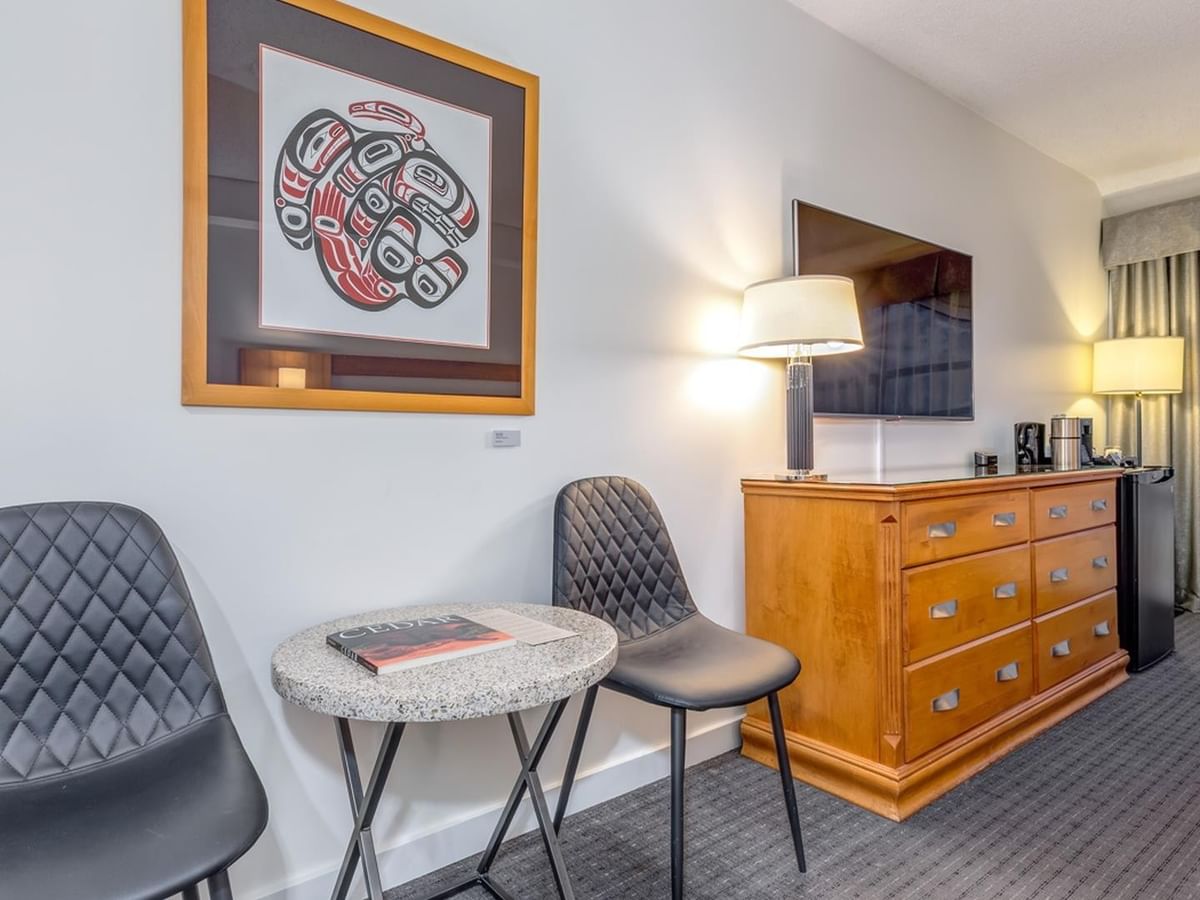 Hotel room with two chairs, small table, dresser, framed Indigenous artwork, TV, and window.