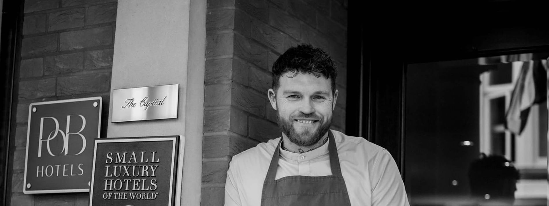 Chef Tom Brown standing by a brick wall featuring hotel signage for The Capital Hotel, Apartments and Townhouse
