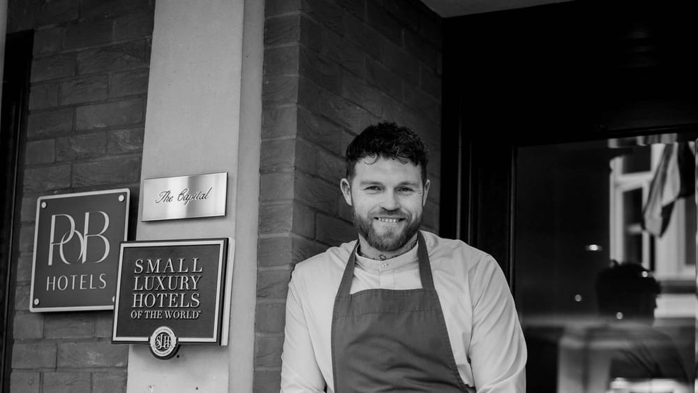 Chef Tom Brown standing by a brick wall featuring hotel signage for The Capital Hotel, Apartments and Townhouse