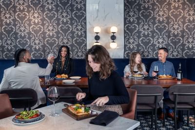 Guests dining with wine at marble tables by blue banquette seating under glowing wall lights at Hotel Saint Clair