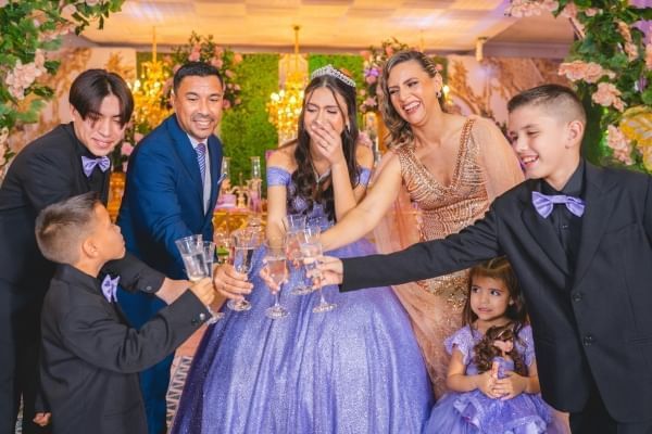 A teen girl in a large purple dress and crown laughs, surrounded by family who are all toasting in glass flutes. 
