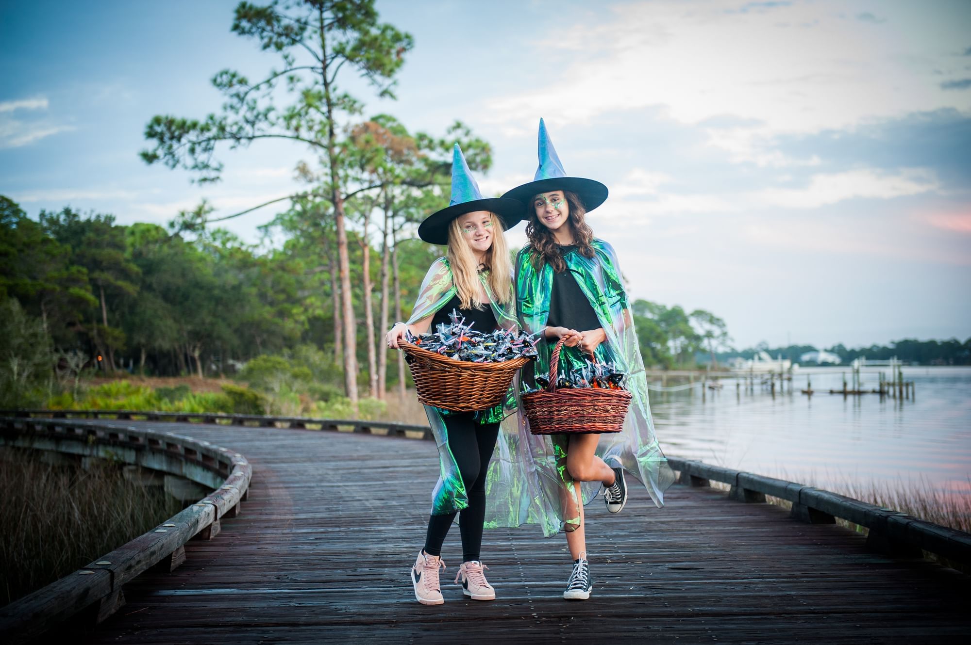 Two women dressed as witches with baskets and capes run down a wooden boardwalk by a lake.