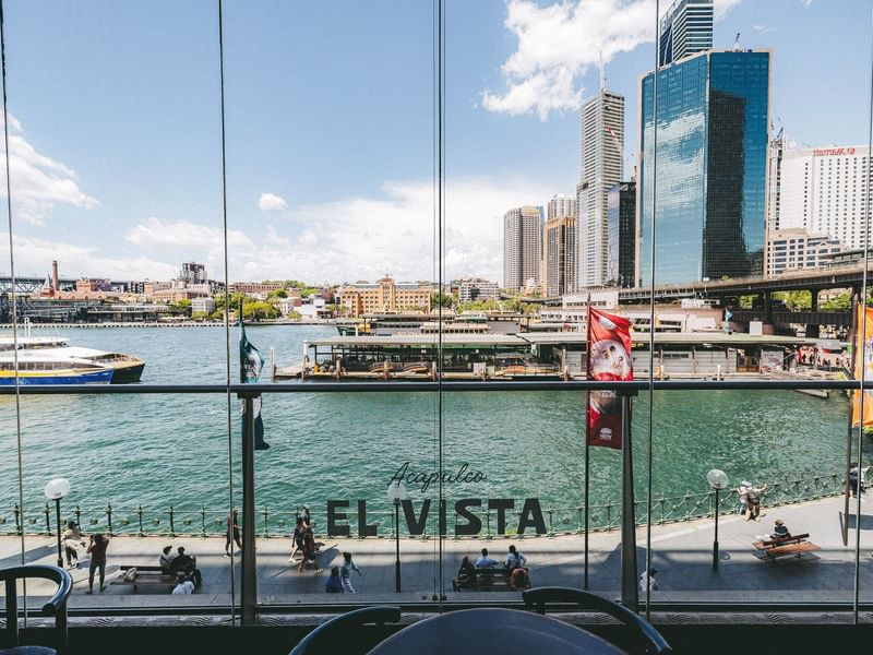 Panoramic harbour view through glass windows with people walking on the waterfront at Pullman Quay Grand Sydney Harbour