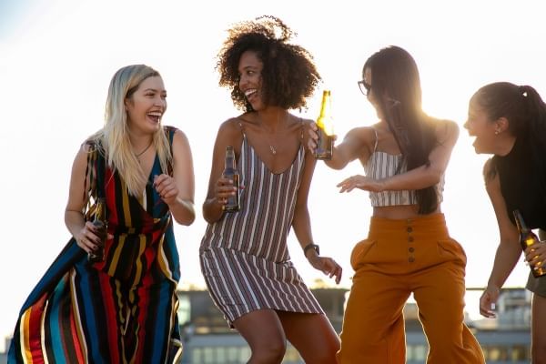 Four ladies holding beer bottles laughing and dancing outdoors. 