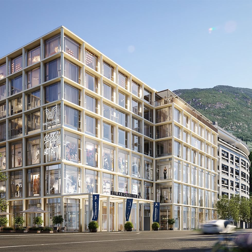 Edificio moderno con vetrine, piante, auto in movimento e montagna sullo sfondo.