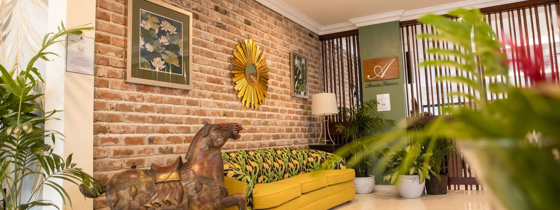 Wooden horse statue by a mustard sofa under framed art and a sunburst mirror on a brick wall at Courtleigh Hotel & Suites