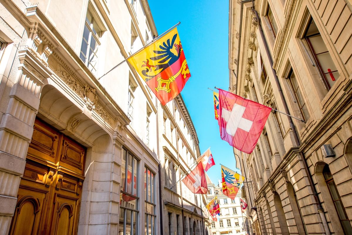 Swiss and regional flags hanging by stone buildings under a bright blue sky near Warwick Geneva