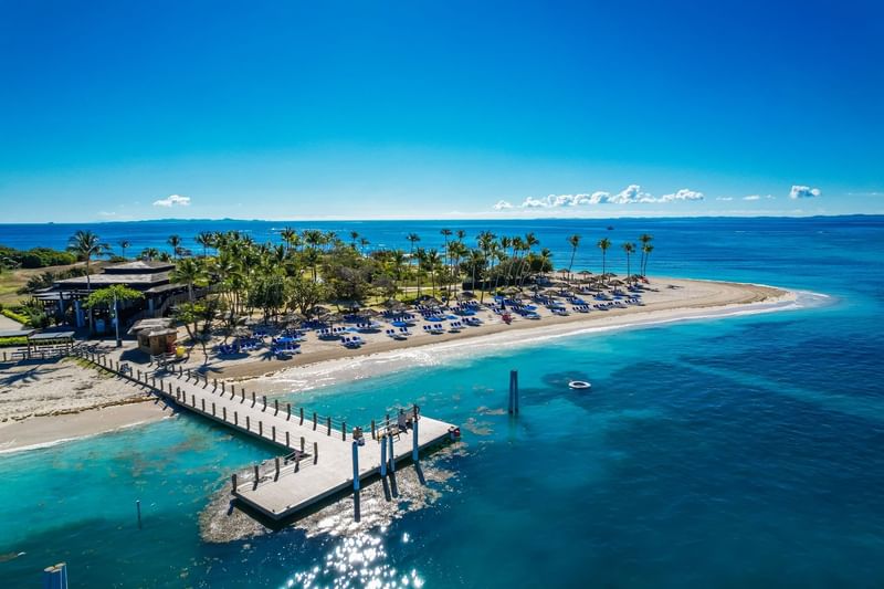 Aerial view of El Conquistador Resort beach with palm trees, lounge chairs, & a wooden pier extending into calm ocean waters