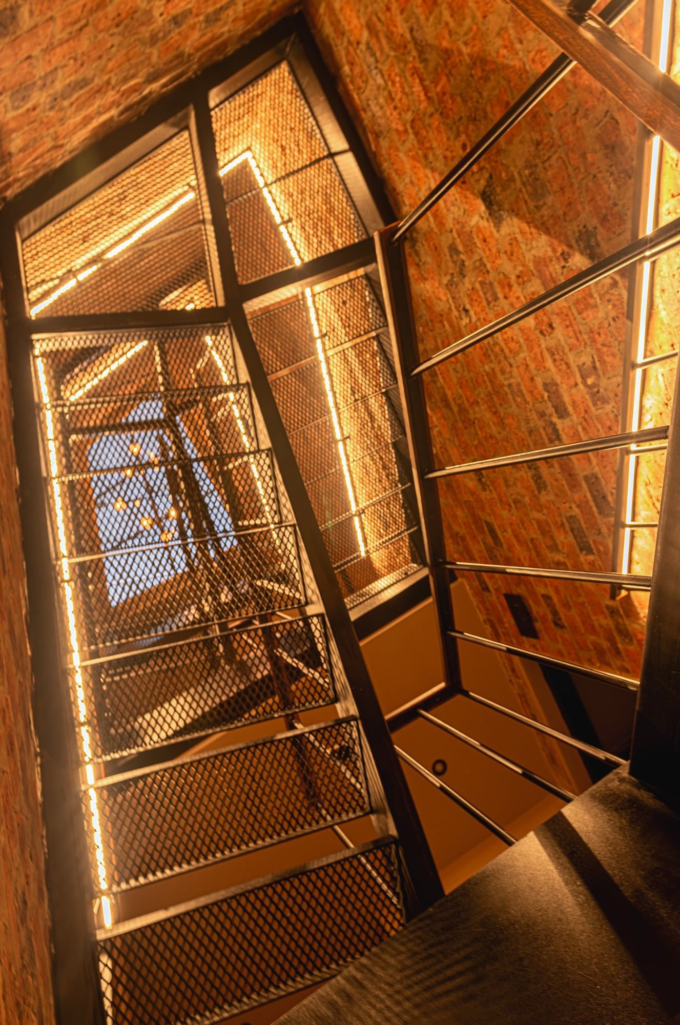 Mesh metal stairs by a red brick wall under linear LED lights and a high ceiling at 5Q House Chapinero