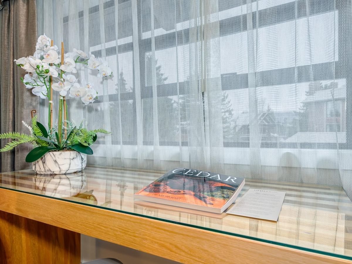 A desk with a book, a vase of white flowers, and a chair by a window.