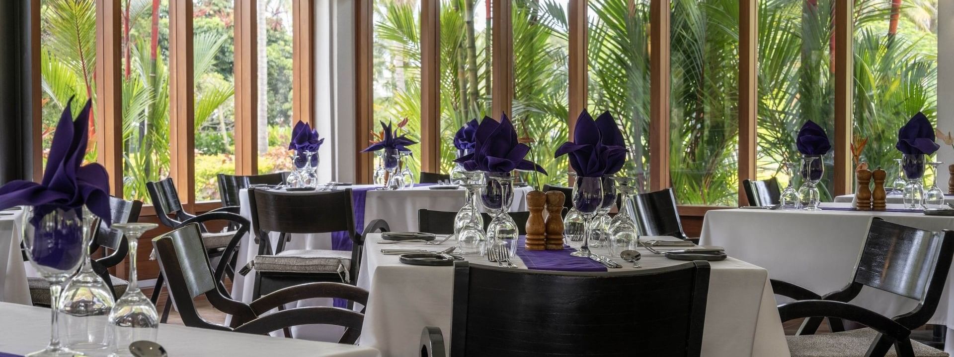 Dining tables with purple napkins by large windows under a white ceiling in Talei Restaurant at The Naviti Resort - Fiji