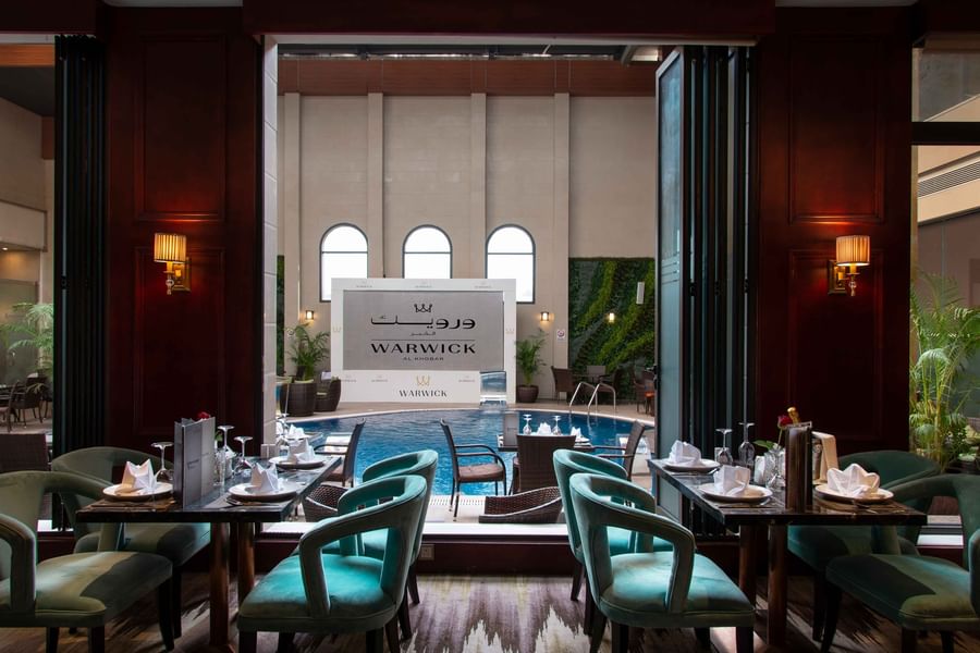 Dining tables with teal chairs overlooking an indoor pool area in Cigar Lounge at Warwick Al Khobar