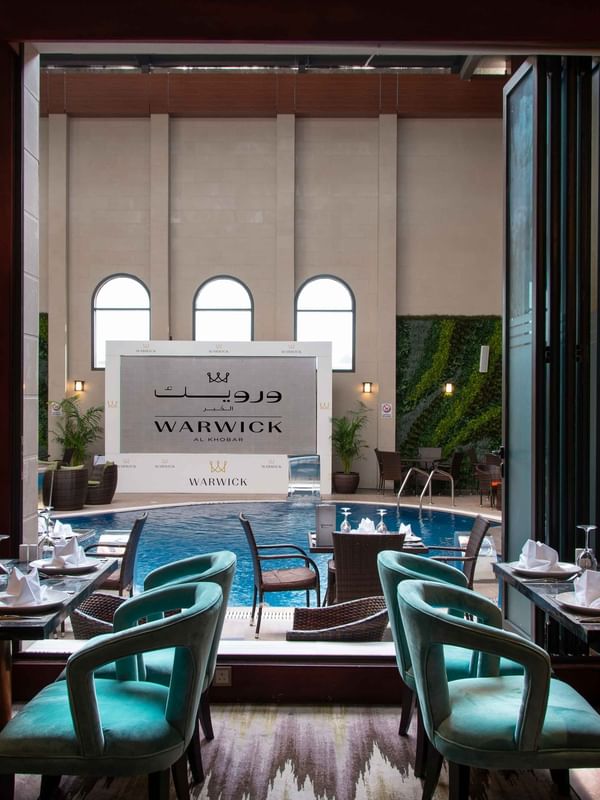 Dining tables with teal chairs overlooking an indoor pool area in Cigar Lounge at Warwick Al Khobar