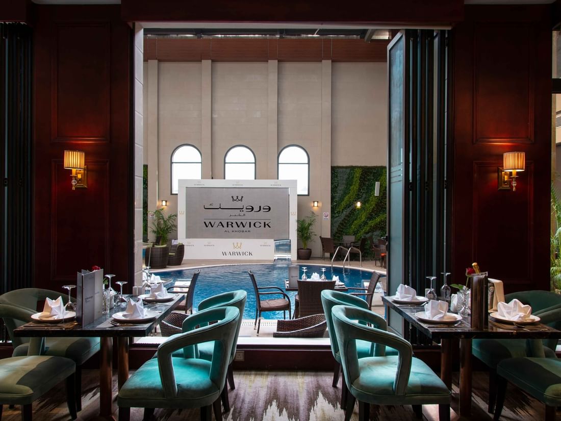Dining tables with teal chairs overlooking an indoor pool area in Cigar Lounge at Warwick Al Khobar