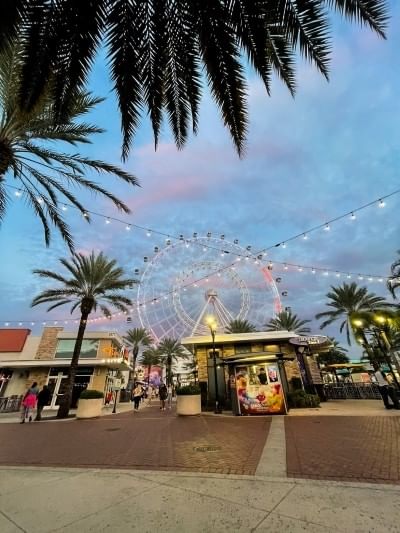A shopping plaza at twilight with a large glowing ferris wheel flanked by palm trees in the distance.