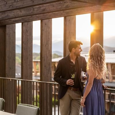 Mann und Frau auf einer Holzterrasse mit Getränken bei Sonnenuntergang, umgeben von Bergen.
