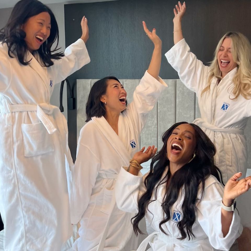 Four women in white robes laughing together on and around a white bed during a relaxing spa getaway at Hotel X Toronto