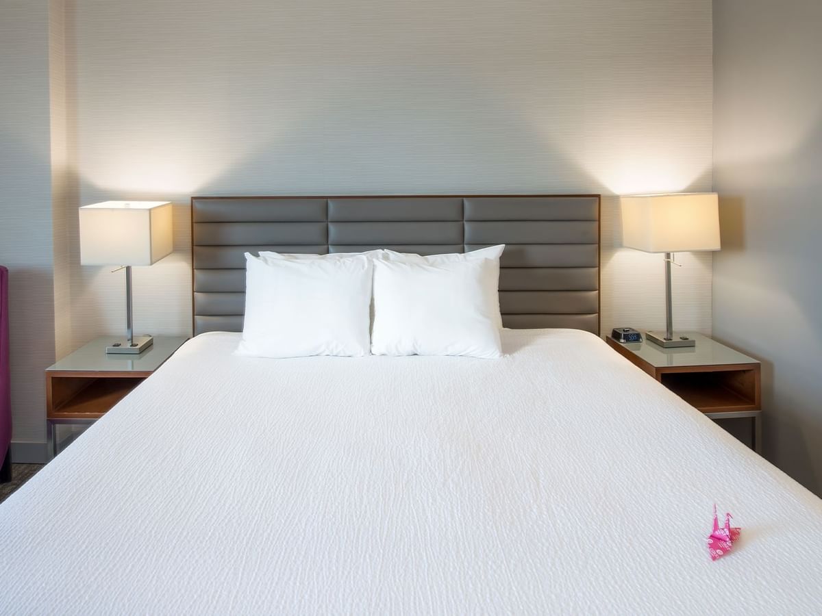A hotel room with a white bed, two pillows, two lamps, a purple chair, and a painting.