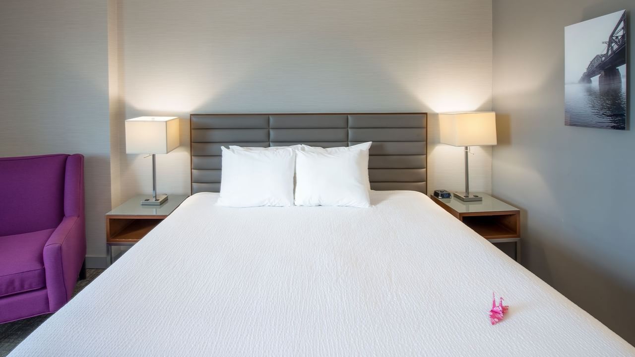 A hotel room with a white bed, two pillows, two lamps, a purple chair, and a painting.
