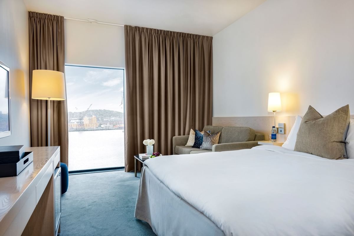 Hotellrum med stor säng, fåtölj och stort fönster med utsikt över vatten.