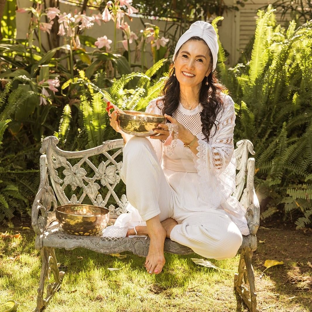 Woman in white attire with head wrap holding golden bowl on garden bench surrounded by lush ferns and pink flowers.