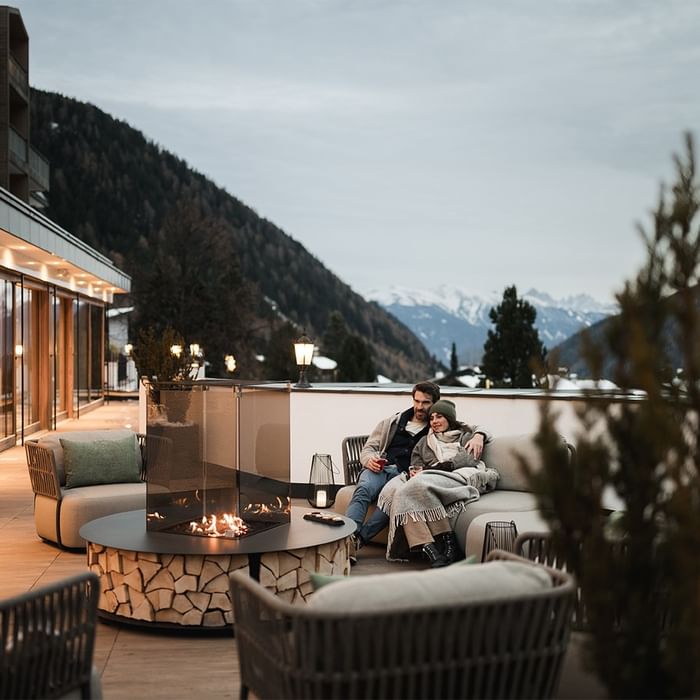 Coppia che si rilassa su una terrazza con vista sulle montagne e camino esterno at Falkensteiner Group