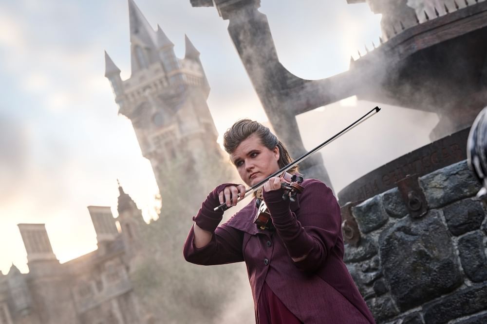 A violin player in a dark purple overcoat surrounded by fog with a creepy tower rising in the background.
