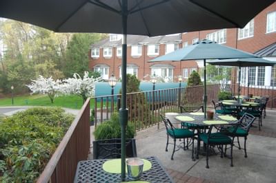 Outdoor patio with black metal tables and green umbrellas beside a brick building and lush trees at The Simsbury Inn