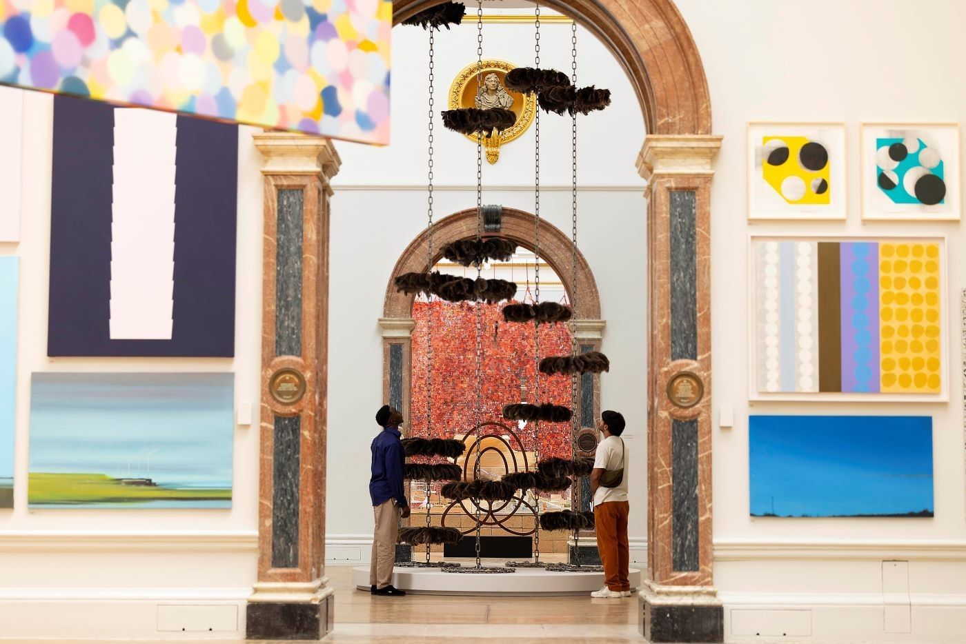 Dois visitantes observam obras de arte contemporânea através de um arco decorado com pinturas abstratas coloridas na Royal Academy of Arts.