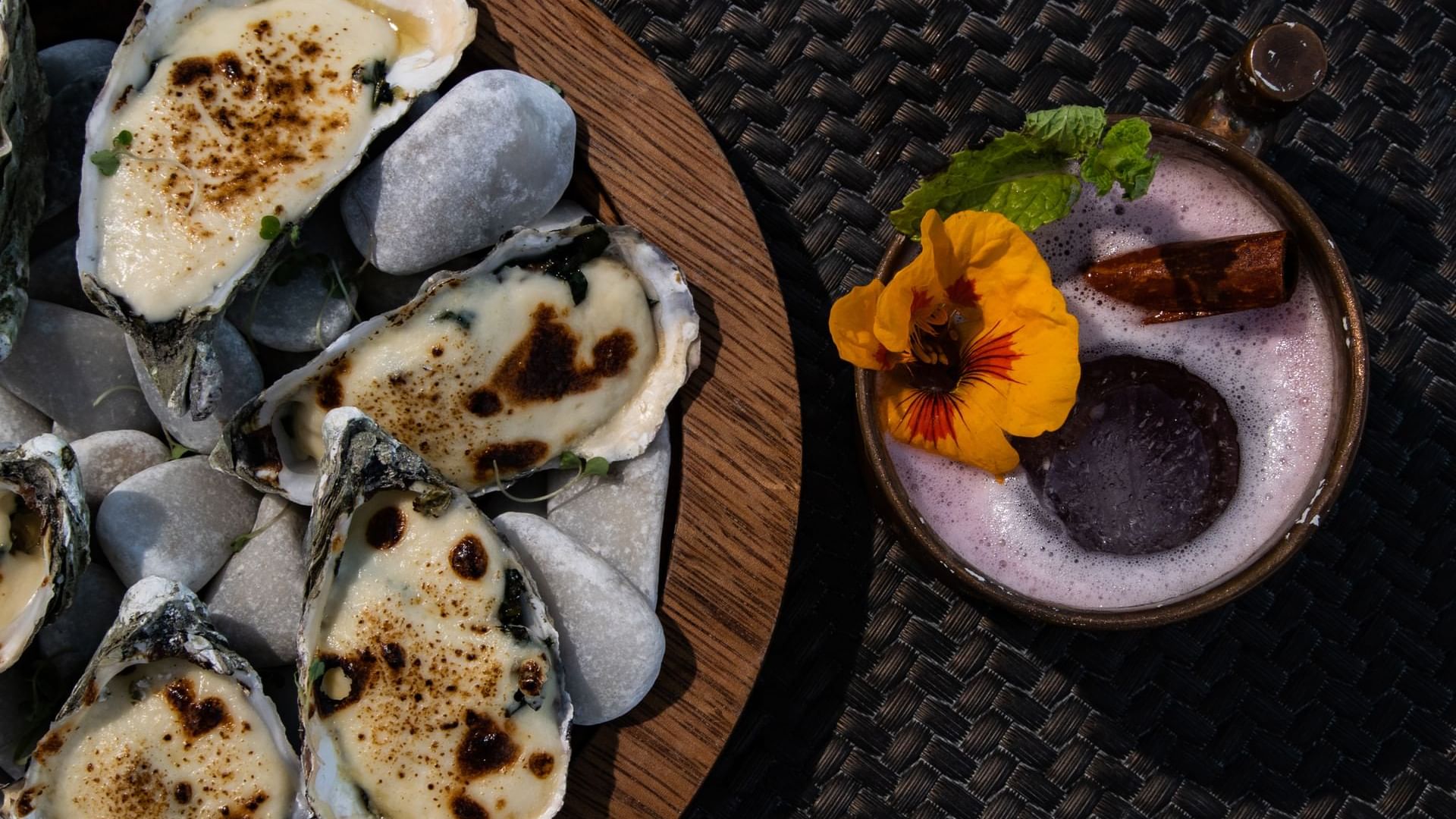 Plato de ostiones horneados sobre piedras, junto a un cóctel festivo en De Cortez Grill House de Hacienda del Mar Los Cabos