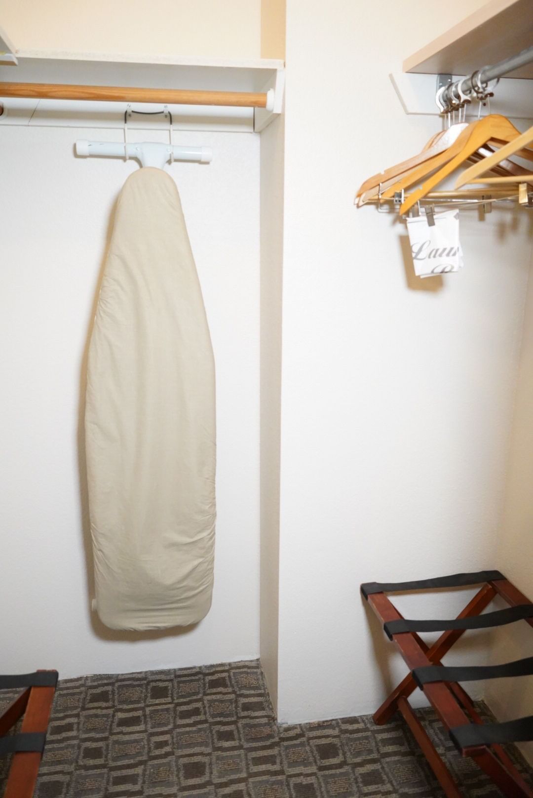 A hotel closet with an ironing board, wooden hangers, and a luggage rack.