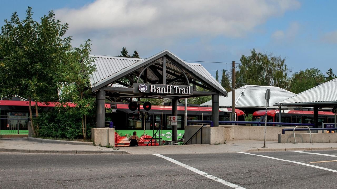 Covered entrance to Banff Trail CTrain Station with red trains visible on the platform behind green trees under a cloudy sky.